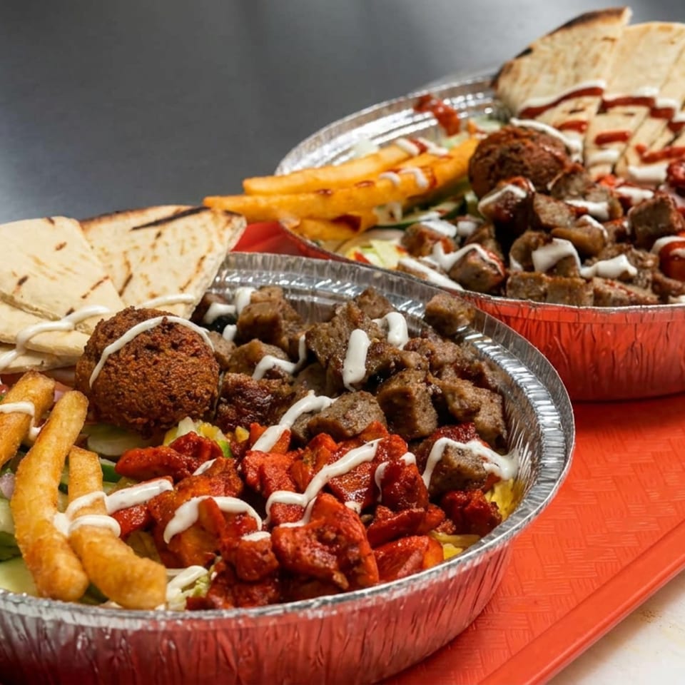 Two foil trays of Middle Eastern meals with grilled meat, falafel, fries, pita bread, salad, and drizzled sauces on a red tray.
