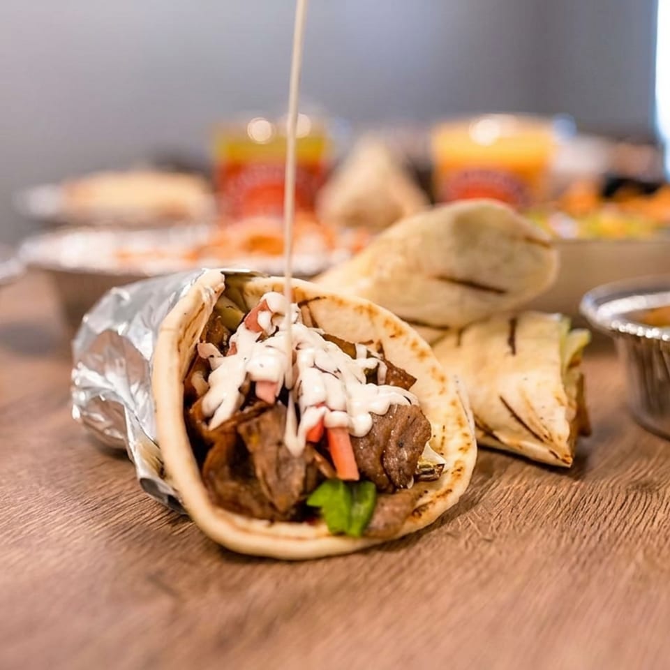 Grilled meat pita wrap topped with creamy sauce and vegetables, surrounded by additional wraps and foil trays of food.