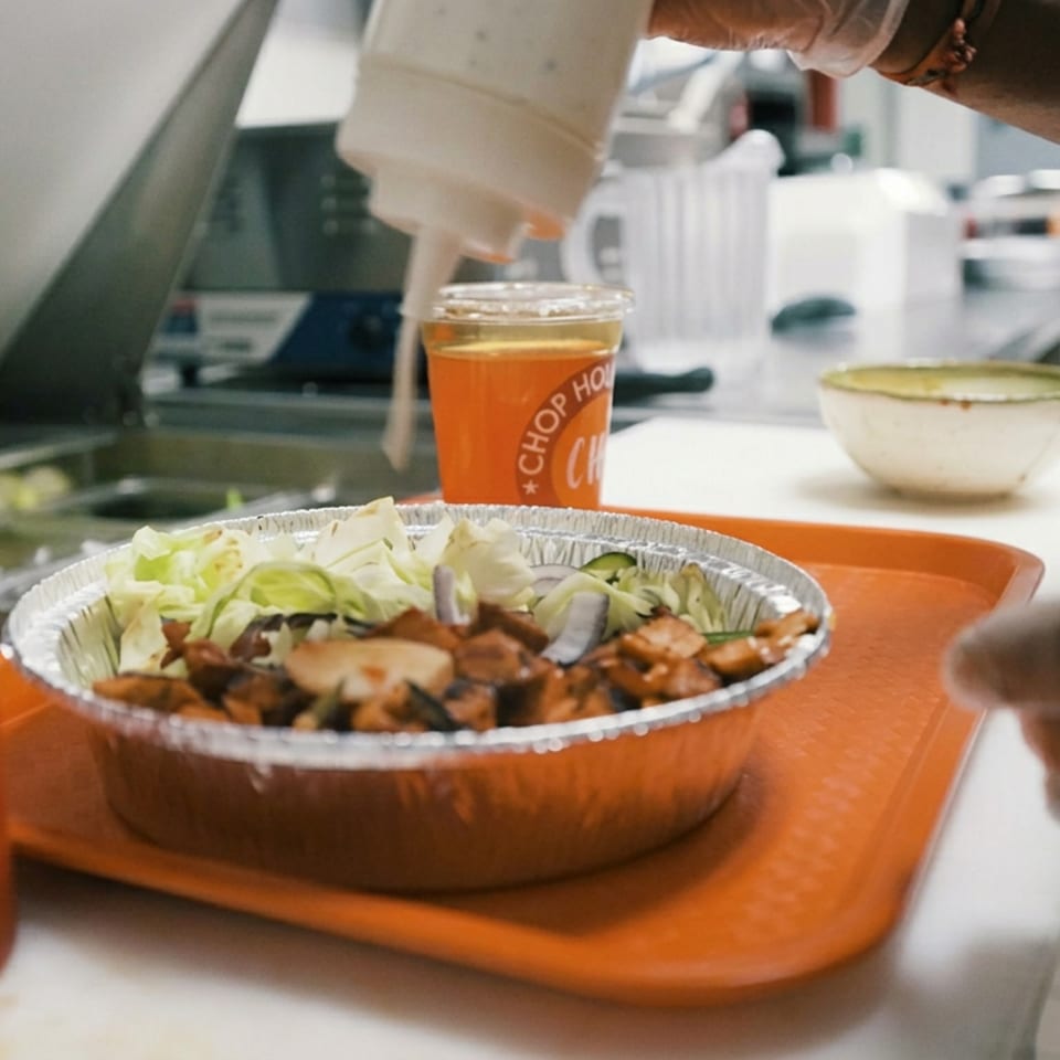 Tray holding a foil bowl of grilled meat, lettuce, and onions, with sauce being added beside an orange drink and condiments.