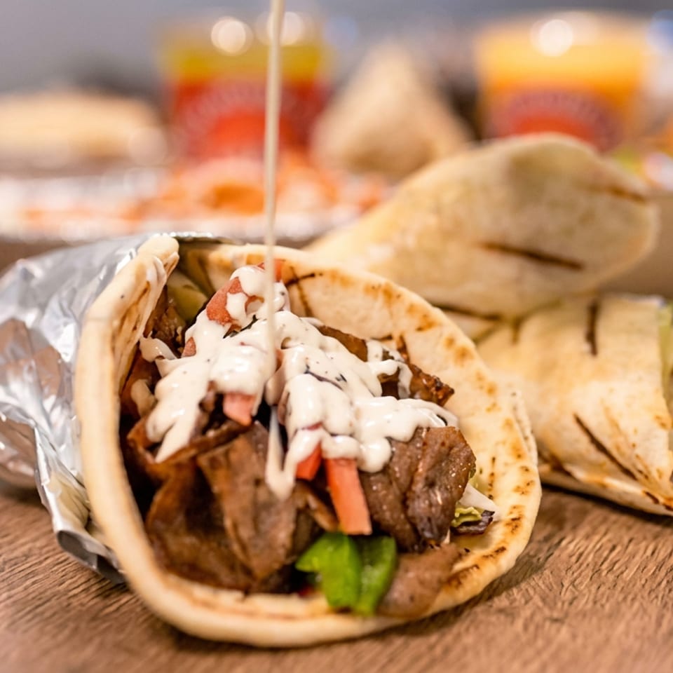 Open pita sandwich filled with grilled meat, vegetables, and creamy sauce, alongside wrapped pita sandwiches and side dishes on a wooden table.