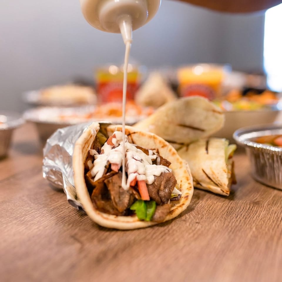 Gyro pita topped with creamy sauce, vegetables, and grilled meat, surrounded by plated Mediterranean dishes on a wooden table.
